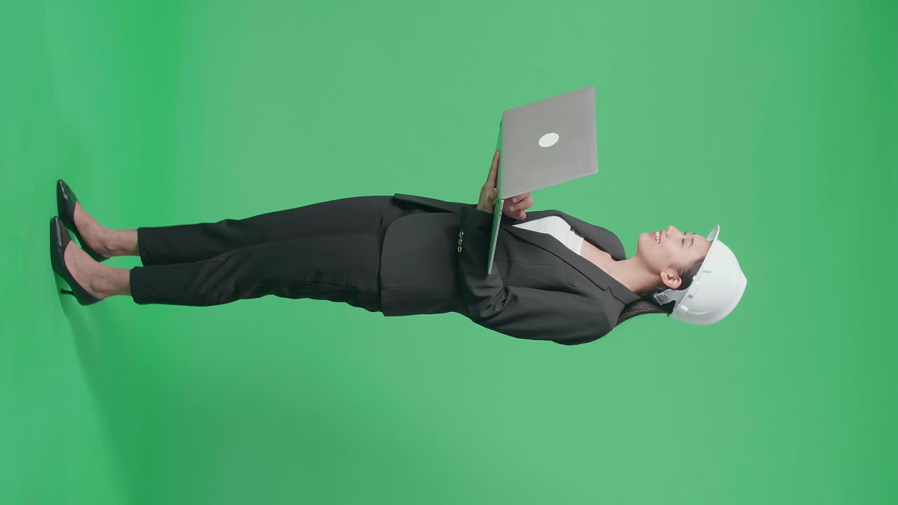 Full Body Side View Of Smiling Asian Female Engineer Looking Around And Shaking Her Head While Using Laptop In The Green Screen Studio
