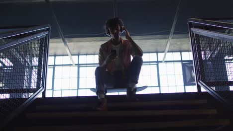 african american man wearing headphones using smartphone while sitting on skateboard