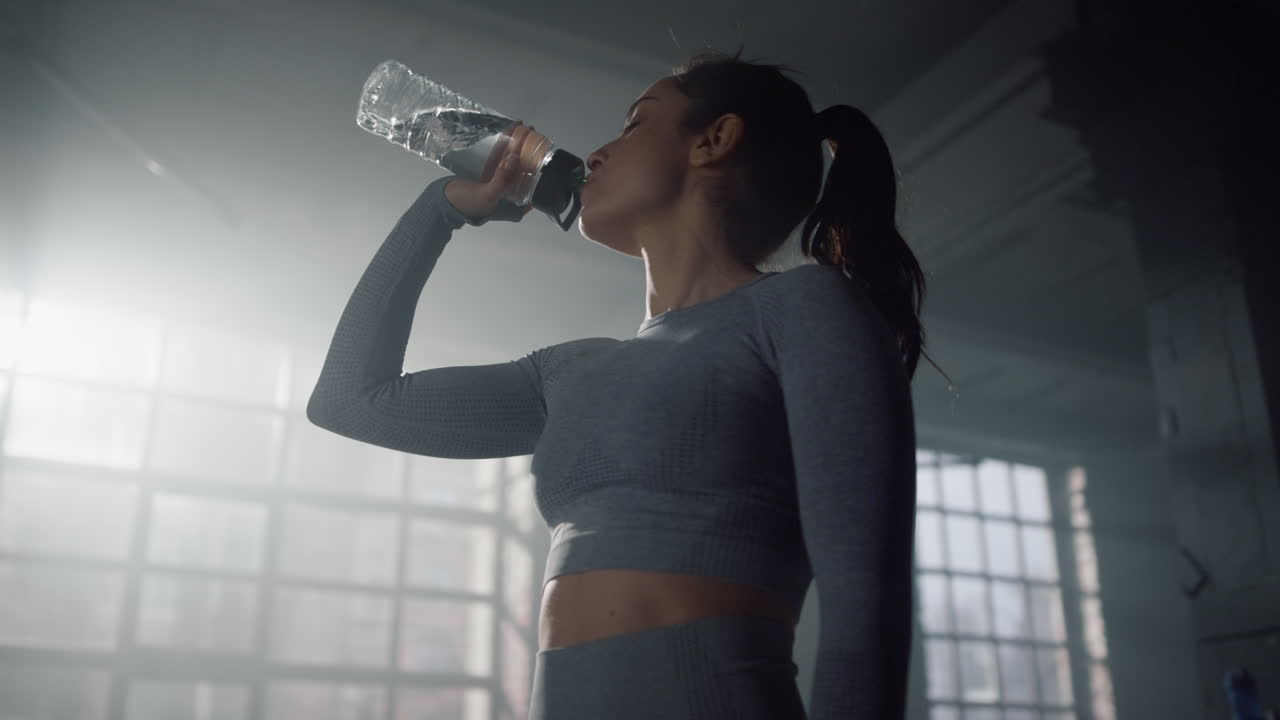 Sportswoman drinking water in gym. Girl taking rest after fitness workout
