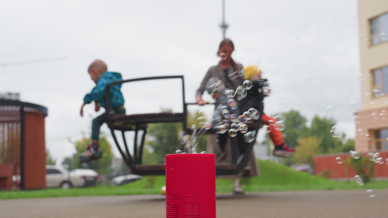 Foreground red bubble machine blows soap bubbles while woman rotates children on merry go round in clockwise motion, blur background with pavilion, crane, green lawn, overcast sky