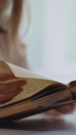 una mujer rubia da la vuelta a las páginas de un libro antiguo sentada cerca de una computadora portátil en un fondo borroso. una estudiante lee información antes de los exámenes en primer plano