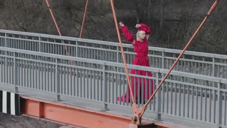 Eine-Wunderschöne-Drohnenaufnahme-Eines-Models-Im-Roten-Kleid,-Das-Auf-Einer-Brücke-Posiert-Und-Tanzt