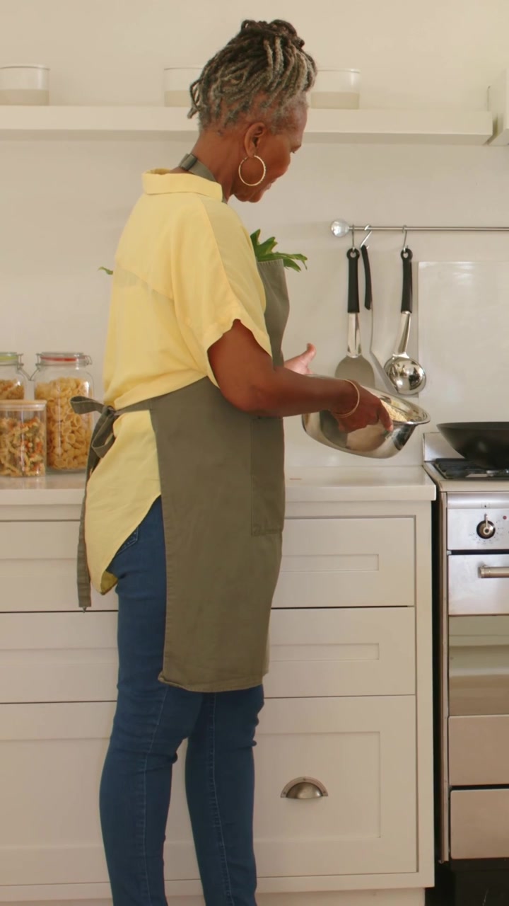 Video verticale: Vedendo la padella, donna con grembiule verde che versa sposta la ciotola per mescolare verso il fornello per cucinare.