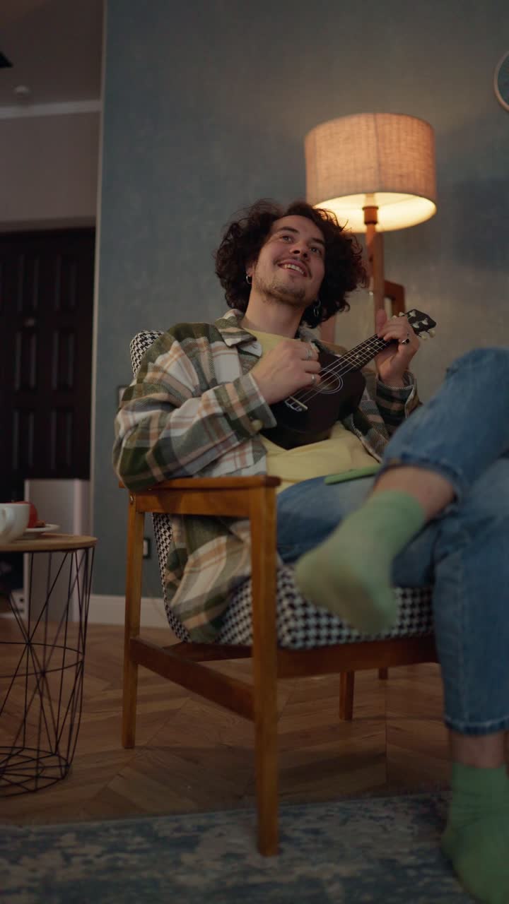 video vertical de un feliz chico moreno con cabello rizado y bigote en una camisa a cuadros sentado en una silla y tocando el ukulele en casa cerca de la lámpara