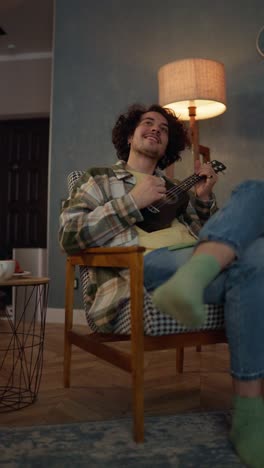 Vertical-video-of-a-happy-brunette-guy-with-curly-hair-and-a-mustache-in-a-checkered-shirt-sitting-on-a-chair-and-playing-the-ukulele-at-home-near-the-lamp