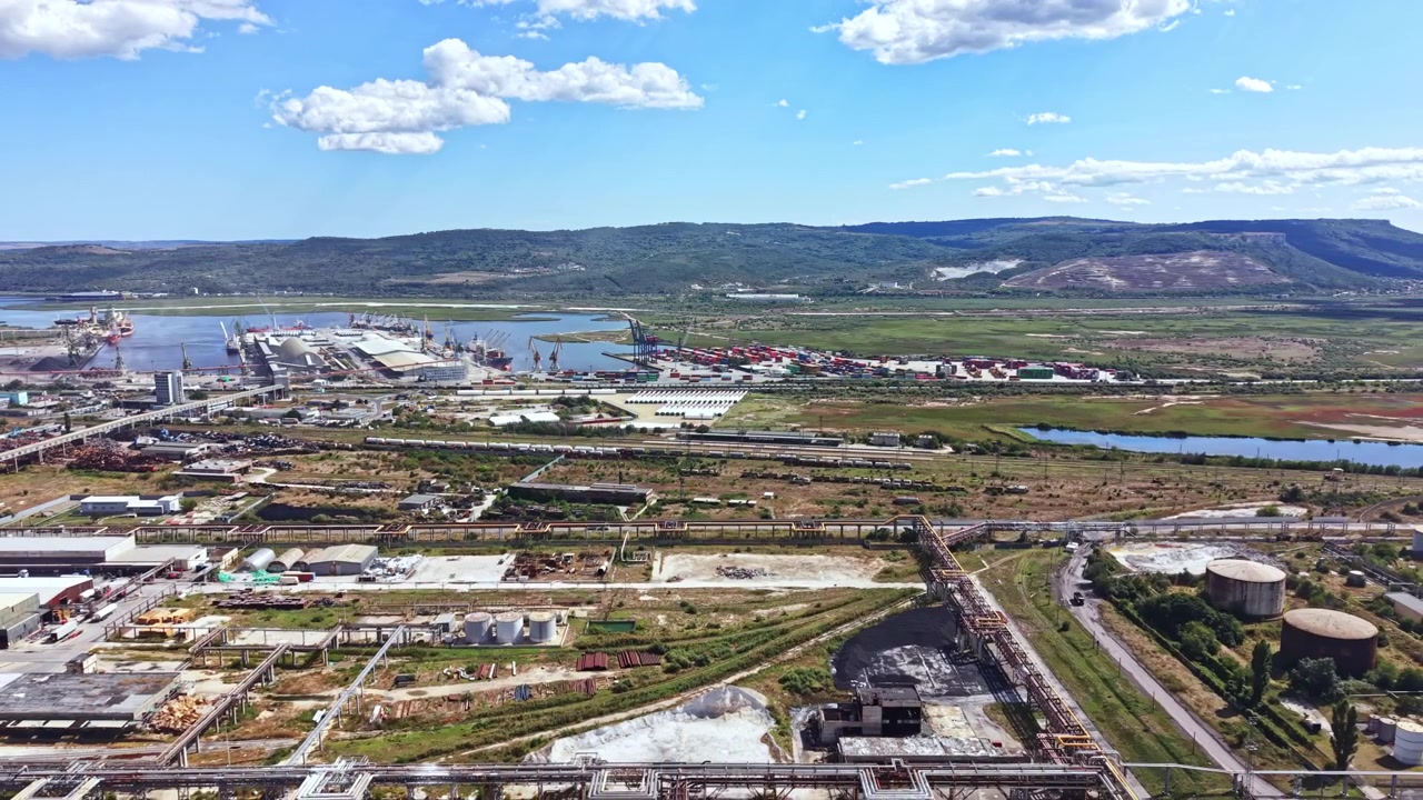 Vista aérea de área industrial com orla, portos de carga, montanhas e campos sob céu azul