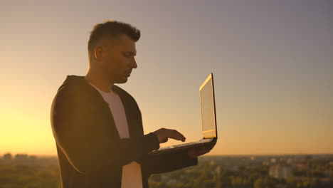 hacker using laptop on rooftop with city view and forex chart. hacking and stats concept. a man at sunset in slow motion writing software code on a laptop