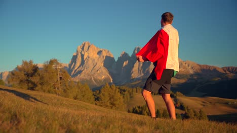 mountains, forest and grass fields with a man holding an italian flag filmed at alpe di siusi in alps, italian dolomites filmed at sunset