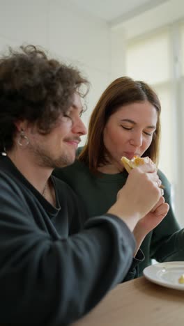 Vertikales-Video-Eines-Glücklichen-Brünetten-Mädchens,-Das-In-Ein-Sandwich-Beißt,-Das-Ihr-Freund-Ihr-Morgens-Beim-Frühstück-Gibt