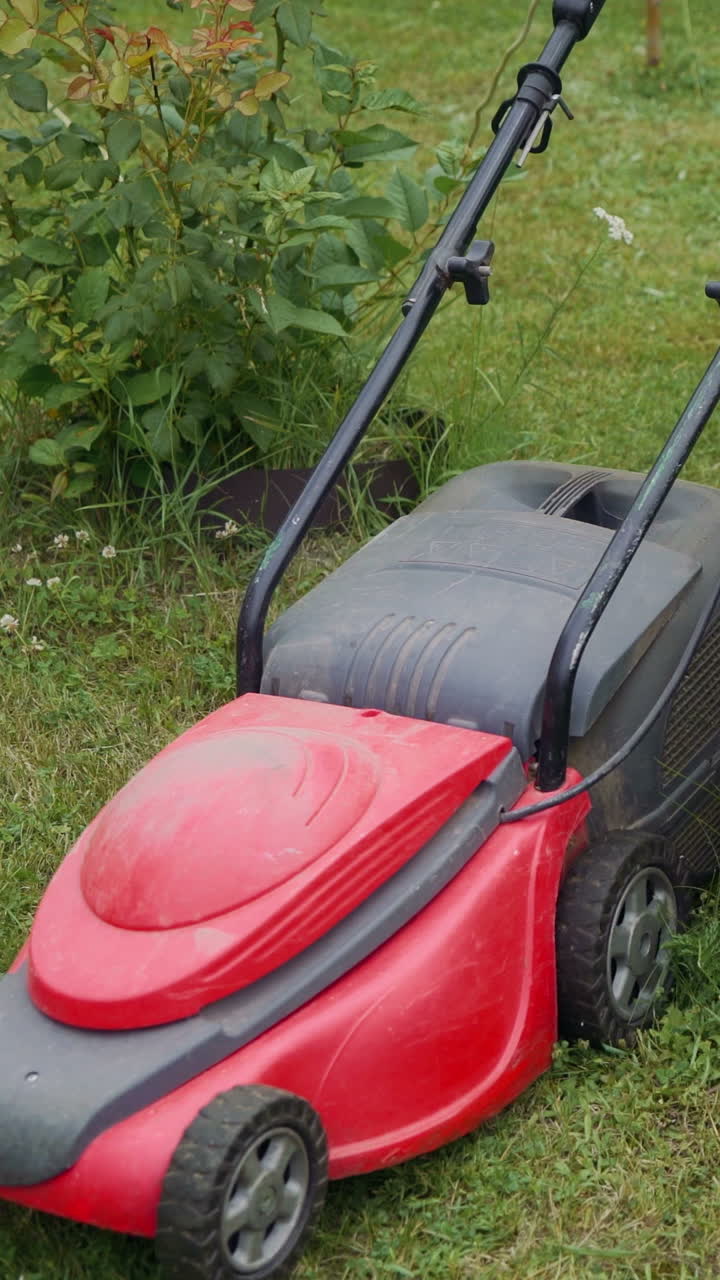 Young woman is mowing lawn with lawn mower in her back yard. Electric lawn mower cuts the grass.. Vertical video