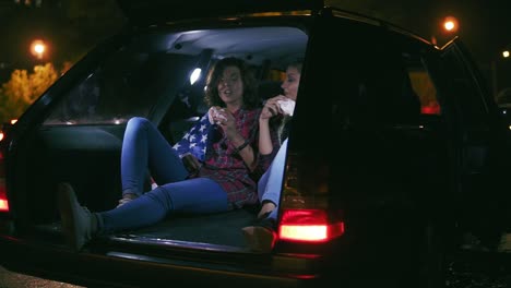 Two-Handsome-Women-Sitting-In-The-Open-Trunk-Of-The-Car,-Talking-And-Eating-Burgers-During-The-Night-In-The-City