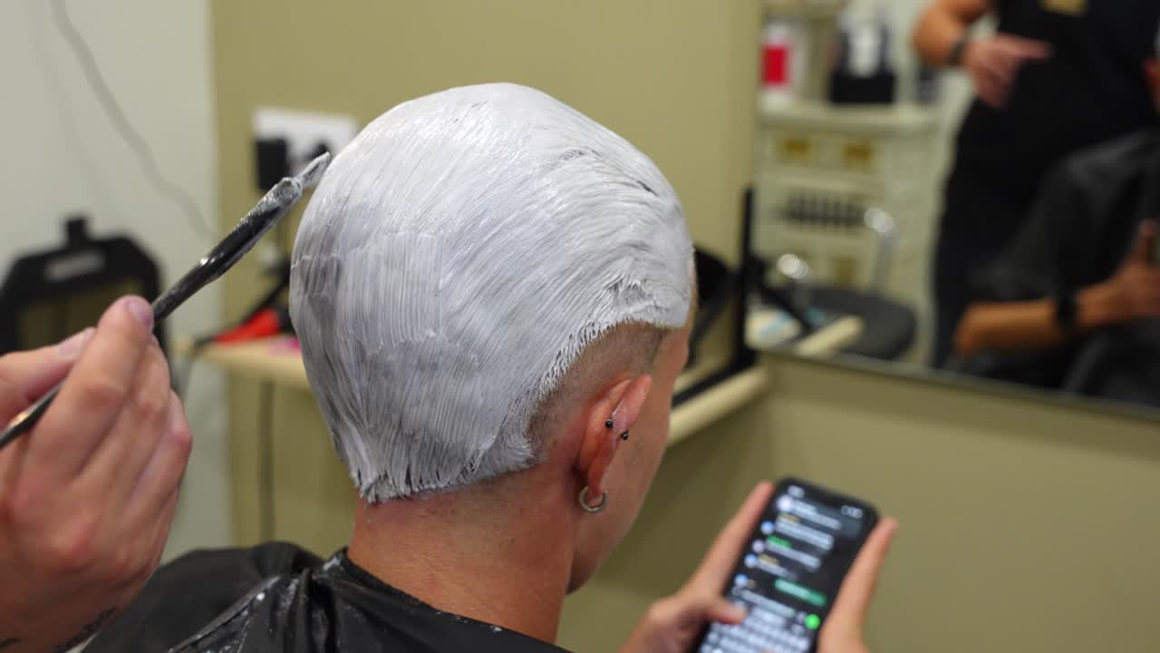 Rear shot of a man in the barber chair using his phone while hairdresser applies bleaching treatment