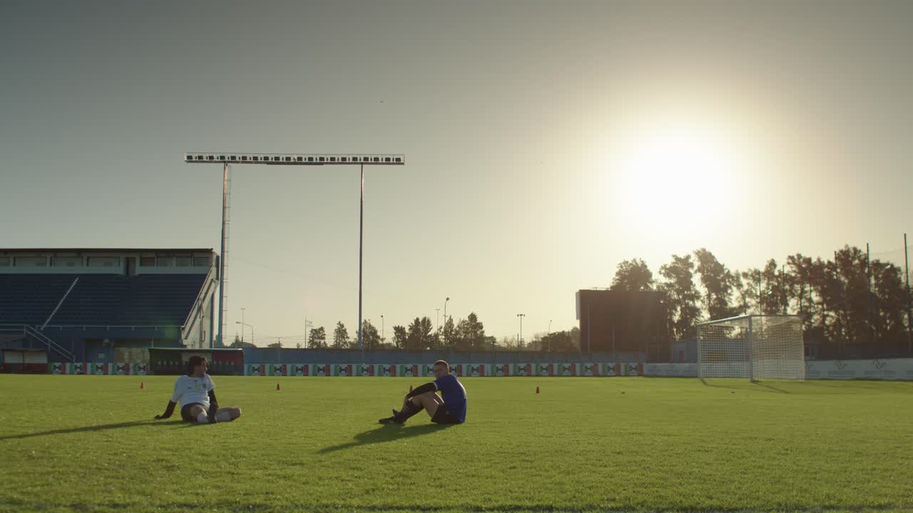 Two football soccer players sit on the field in the late afternoon sun after practice ends
