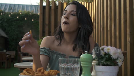 Adorable-woman-eating-curly-fries-in-a-cafe-in-London,-hispanic-latina-millennial-at-the-table-in-a-casual-outfit,-eating-and-having-fun,-smiling-and-enjoying-the-fast-food