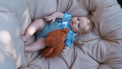 cute caucasian baby boy lies comfortably in egg chair. infant looks at the brown teddy bear in his hands. vertical screen.