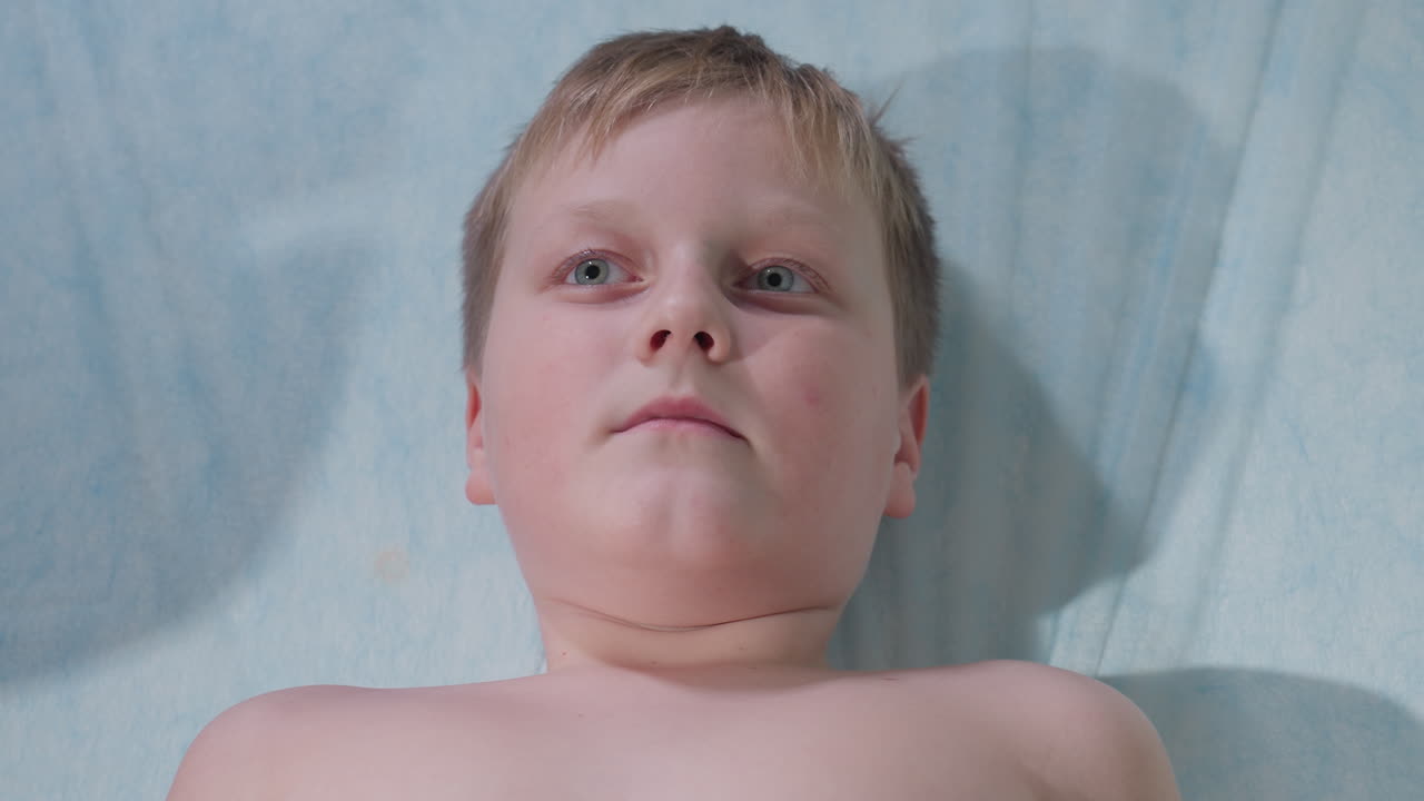 Sick young patient lying barechested on medical bed blinks multiple times, opens and closes mouth gently during examination, portraying subtle discomfort and quiet endurance in clinical setting