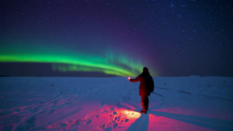 person with red light under vibrant northern lights in snowy landscape