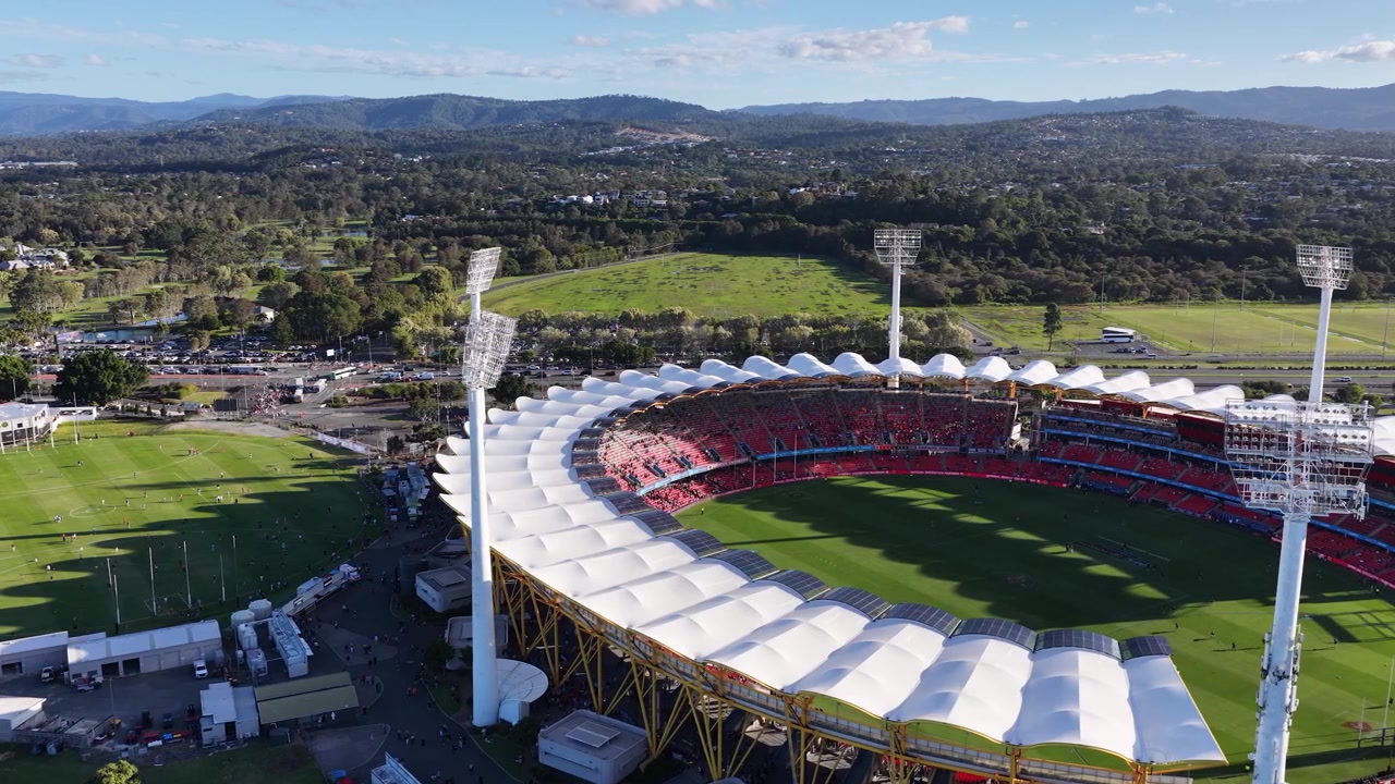 Drone footage pans across a large sports stadium with white roof and surrounding green hills.
