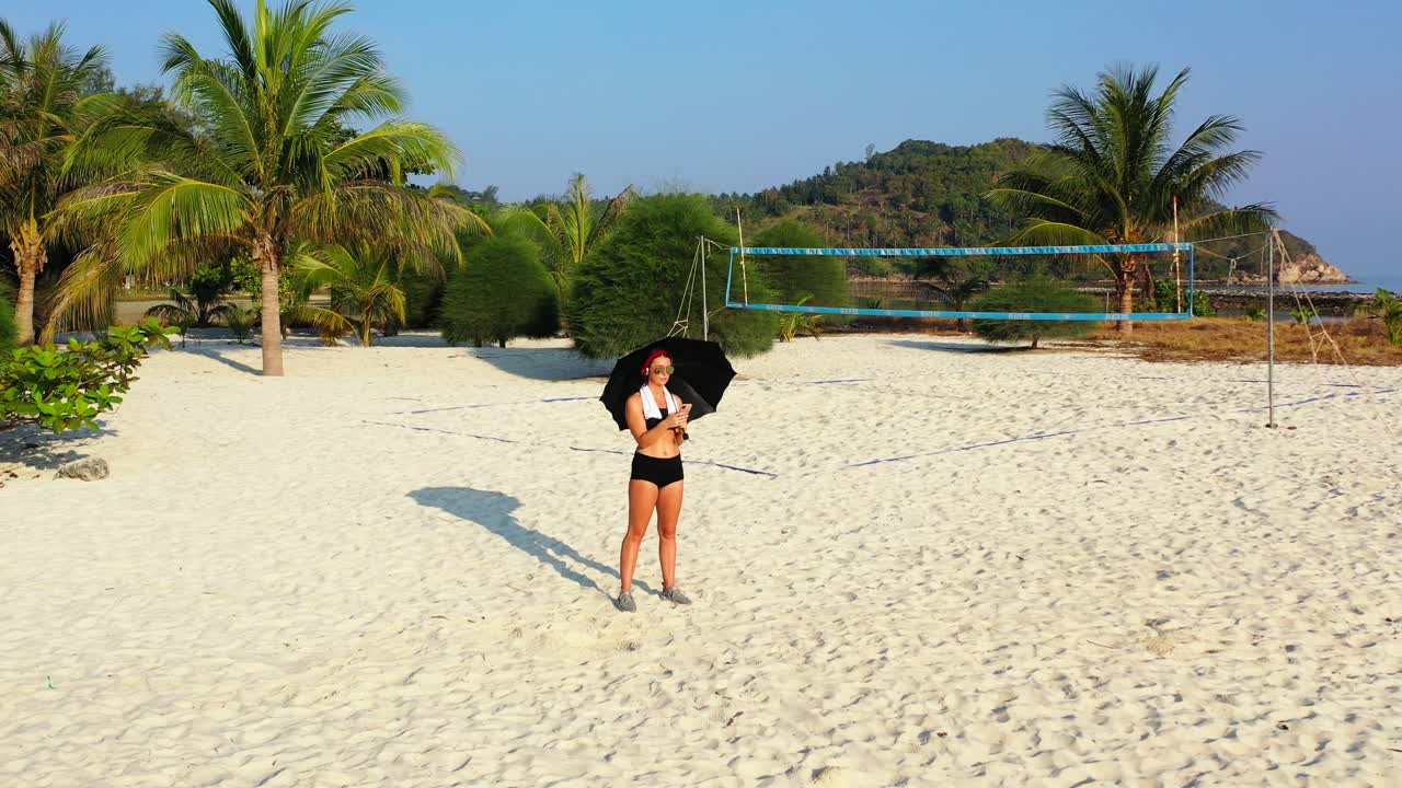 Stylish girl wearing trendy sport fashion clothes with black umbrella and headphones reading smartphone messages on exotic beach in Malaysia
