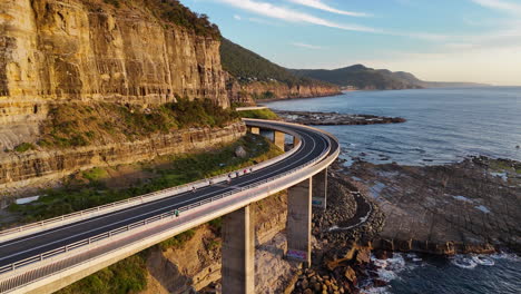 aerial view of the sea cliff bridge, following a group of bicyclists on one of australia’s most scenic oceanfront roads. drone footage.