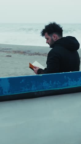 man reading a book sitting on the beach in winter near a boat
