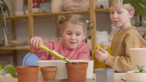 Niña-Rubia-Y-Niño-Rubio-Preparando-El-Suelo-Y-Regando-Una-Olla-Sentada-En-Una-Mesa-Donde-Hay-Plantas-En-Un-Taller-De-Artesanía