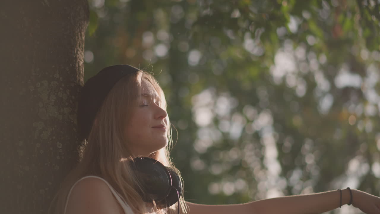Giovane donna con le cuffie che sorseggia una bevanda in un parco soleggiato, appoggiata a un albero con riverbero dell'obiettivo e una calda retroilluminazione, occhi chiusi ed espressione serena, pausa melodica e atmosfera riflessiva nell'inquadratura