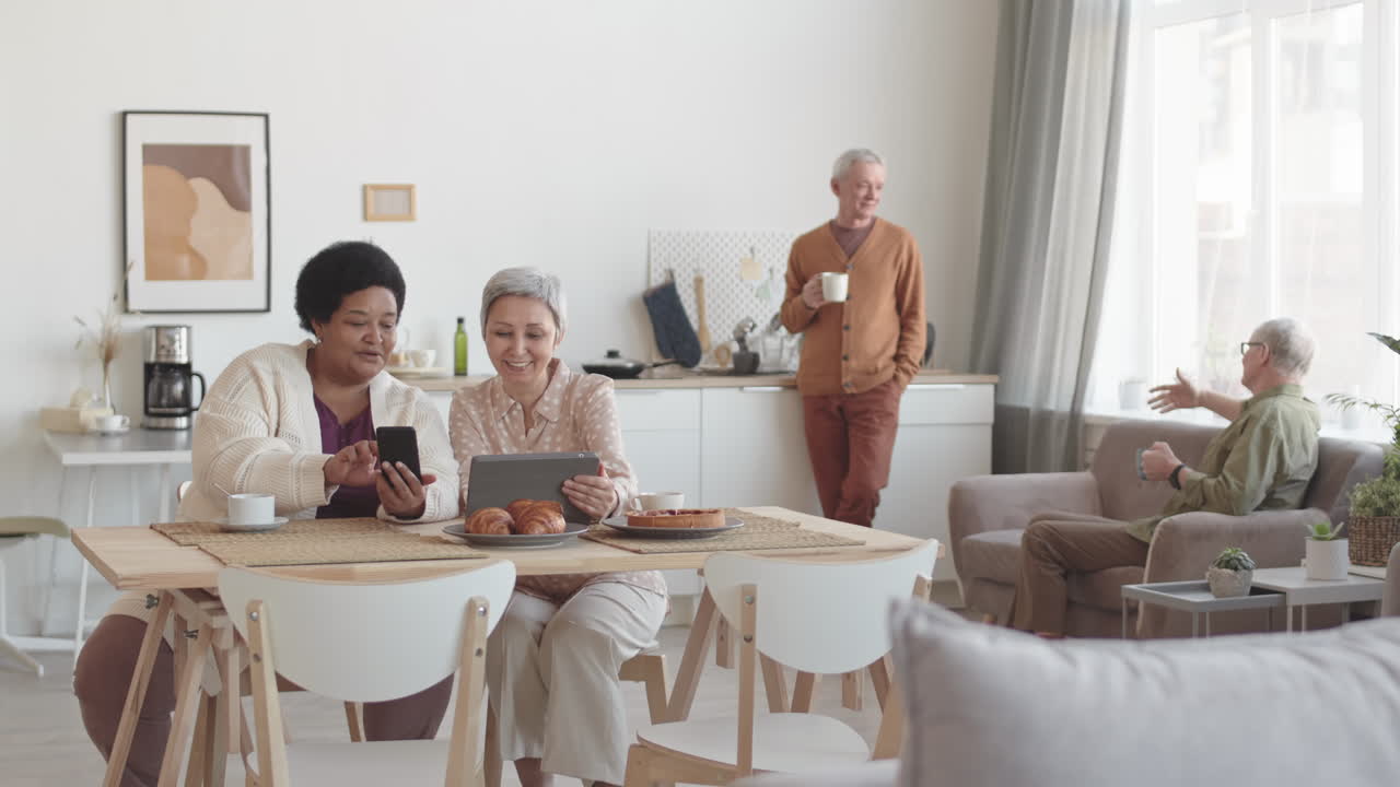 Wide shot of Asian and African mature women sitting at table in room, chatting, having tea with pastries, using tablet computer and smartphone, men communicating on background
