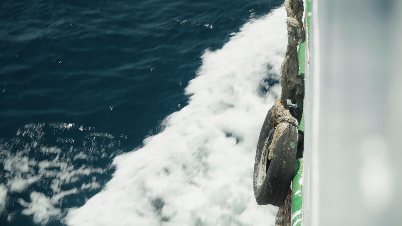 Kolkend kielzog naast bootfender, close-up van turbulent donkerblauw water dat opspat tegen de scheepsreling, schuimende nevel en ritmisch schuim, groene romprand met versleten fender, perspectief aan boord