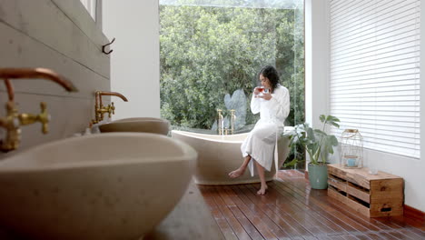 biracial woman wearing bathrobe sitting on bathtub drinking tea at home, slow motion, copy space