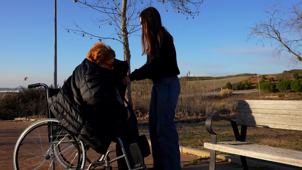 Girl helping grandmother in wheelchair to put on coat in sunny park outdoors