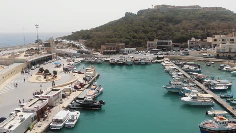 Mgarr-Harbour-with-anchored-vessels-in-the-rugged-coastline-of-the-Gozo-Island-in-Malta---Fly-over-aerial-shot