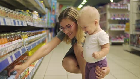 Young-attractive-mother-in-glasses-choosing-baby-food-on-the-shelves-in-the-supermarket-together-with-her-cute-child.-Mom-and