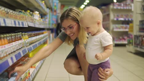 joven madre atractiva con gafas eligiendo comida para bebés en los estantes del supermercado junto con su lindo hijo. mamá y