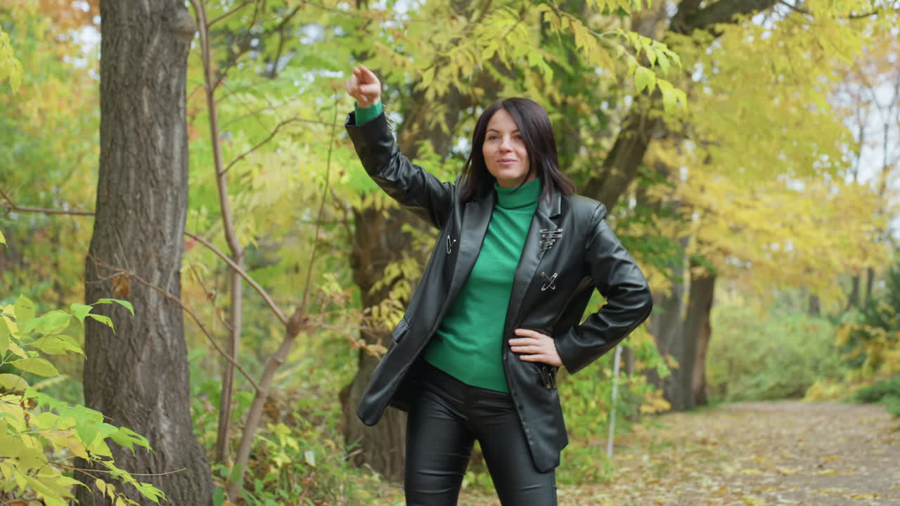 Mulher confiante com jaqueta de couro no parque no outono. Suéter verde sob casaco de couro preto, fazendo poses marcantes ao longo do caminho arborizado. Mão no quadril, gesto apontando, leve virada de cabeça e caminhada relaxada criam