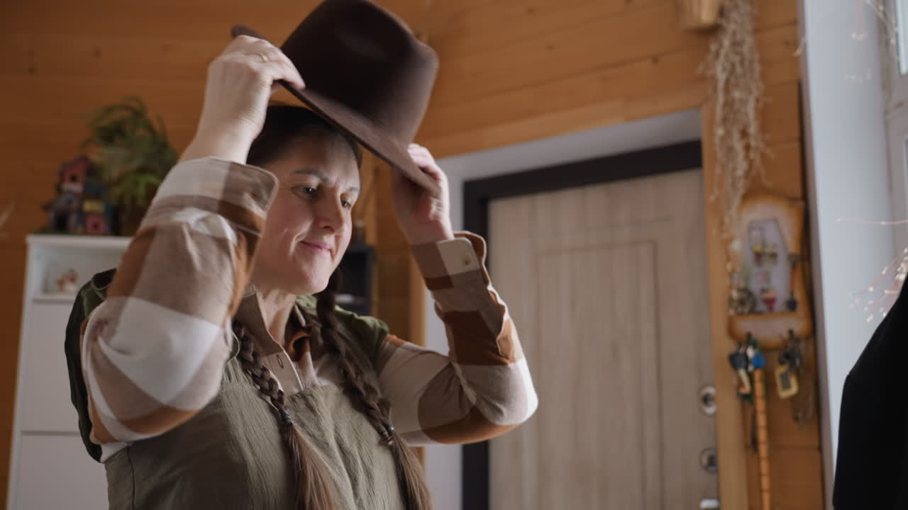 Plant lover with braided hair smiles while grabbing cap from hanger and placing it on her head in wooden room decorated with potted plants and baskets under natural light in cozy indoor setting