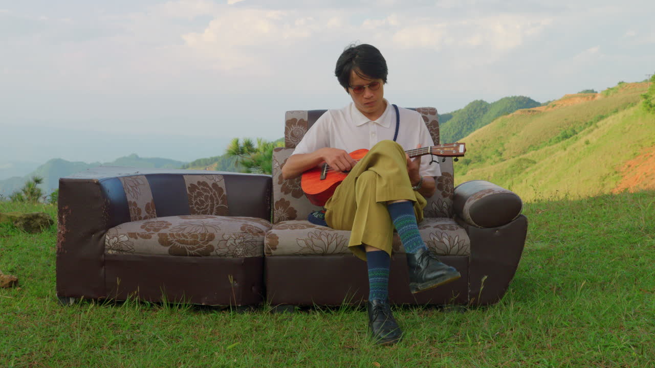 young artist musician playing song ukulele guitar while sitting on couch placed in mountains valley , extravagant artist looking for inspiration