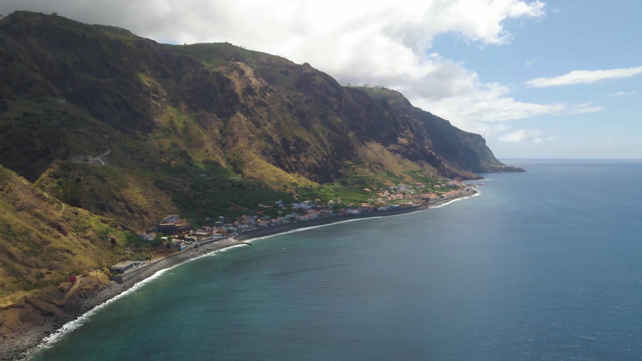 Ripresa drone che vola lungo la costa di Paul do Mar, rivelando ripide scogliere vulcaniche e un piccolo villaggio costiero. Le onde dell'Atlantico si infrangono ritmicamente sulla riva, creando una scena pacifica.
