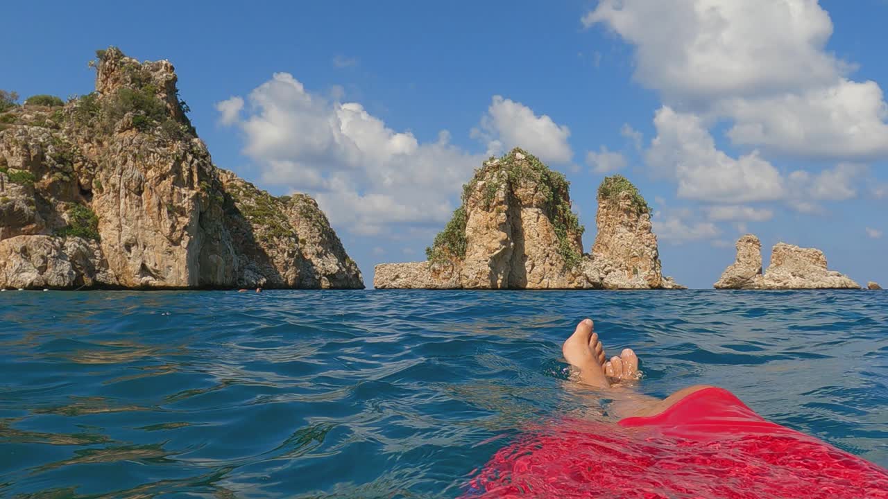 fpv de piernas y pies de hombre relajándose mientras flotan en el agua del mar con pilas de scopello o faraglioni en el fondo