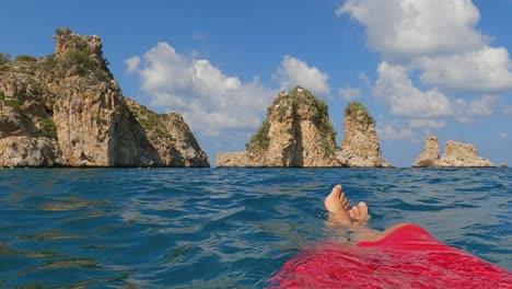 FPV-De-Piernas-Y-Pies-De-Hombre-Relajándose-Mientras-Flota-En-Agua-De-Mar-Con-Pilas-De-Scopello-O-Farallones-En-El-Fondo