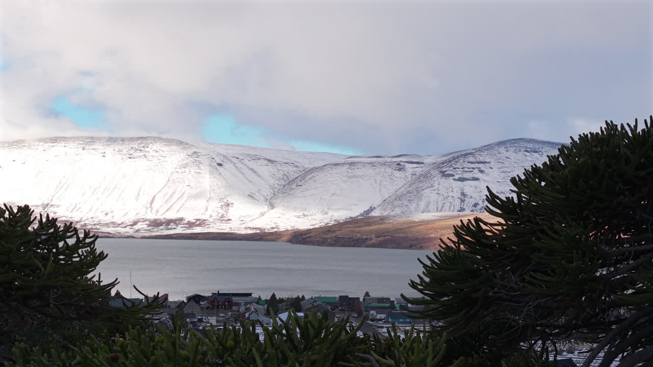 Drone panning shot revealing Caviahue town, snowy hills, and a lake under a cloudy sky in Patagonia, Argentina