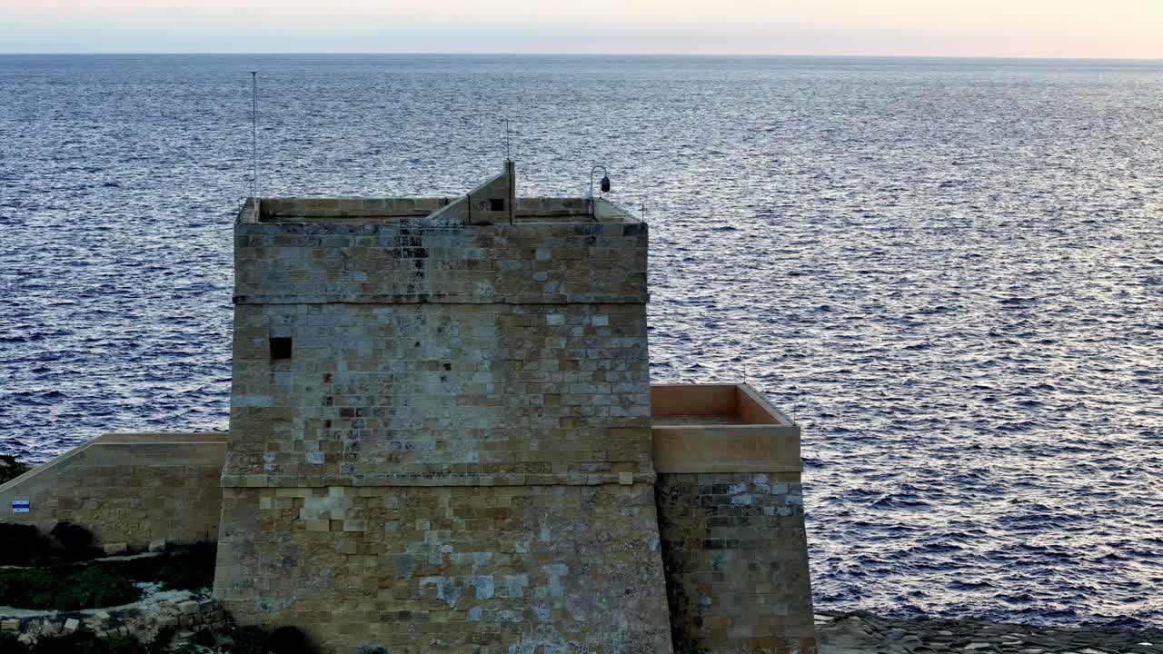 Det eldgamle kalksteinstårnet Dwejra Tower står som en stille vaktpost over det dypblå Middelhavet under en rolig gyllen time på Gozo, Malta, og fanger en majestetisk historisk kystforsvarssilhuett