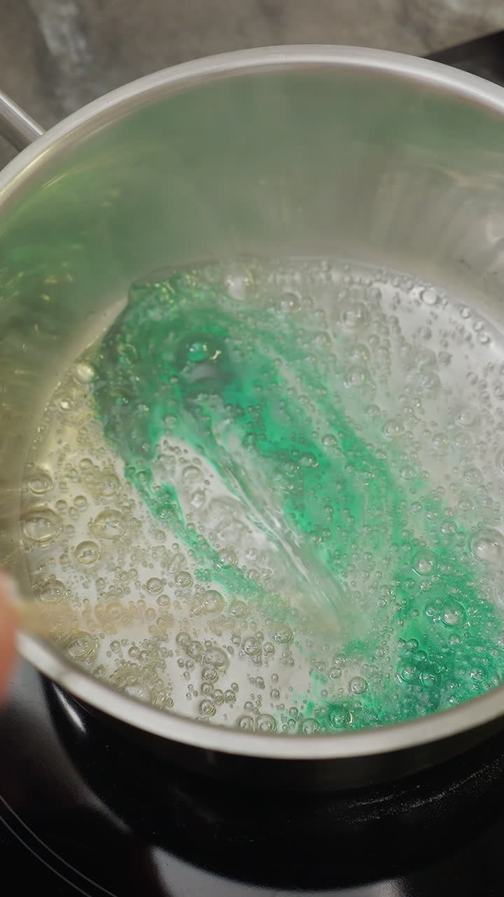 Close-up of person stirring in pot with green colorant, steam rising from liquid, beads of different colors green and golden on wooden tray in background, cooking or food preparation process