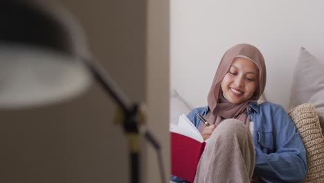 Video-of-smiling-biracial-woman-in-hijab-writing-in-notebook-sitting-in-living-room-at-home