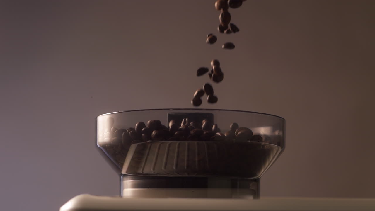 Primo piano al rallentatore di chicchi di caffè tostati che cadono in un contenitore trasparente per macinacaffè da espresso. L'illuminazione calda e soffusa mette in risalto il movimento, la consistenza e la fase di preparazione del caffè speciale
