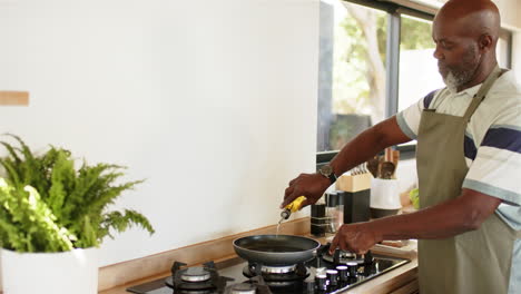 cooking in kitchen, man adding oil to frying pan on stove, wearing apron