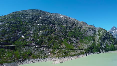 Aerial-drone-backward-moving-shot-over-a-winding-road-along-the-slope-of-Grimselpass-high-mountain-road-in-beautiful-Swiss-Alps,-Switzerland-on-a-sunny-day