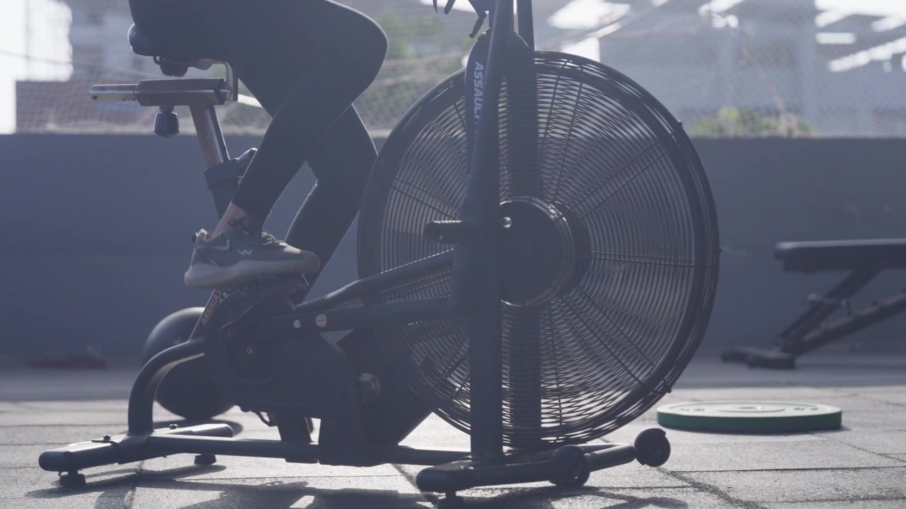 Close-up of the lower body of a South Asian female athlete pedaling an assault air bike during functional fitness workout in a modern indoor fitness studio. Collection: WORKOUTSERIES2026.