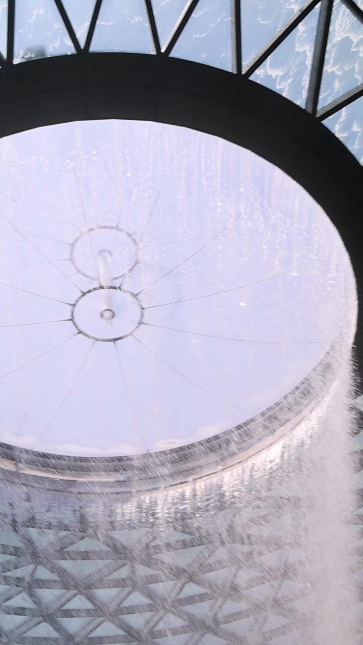 Camera tilts upward from lush indoor trees to reveal a dramatic indoor waterfall cascading from a geometric glass dome ceiling in bright daylight