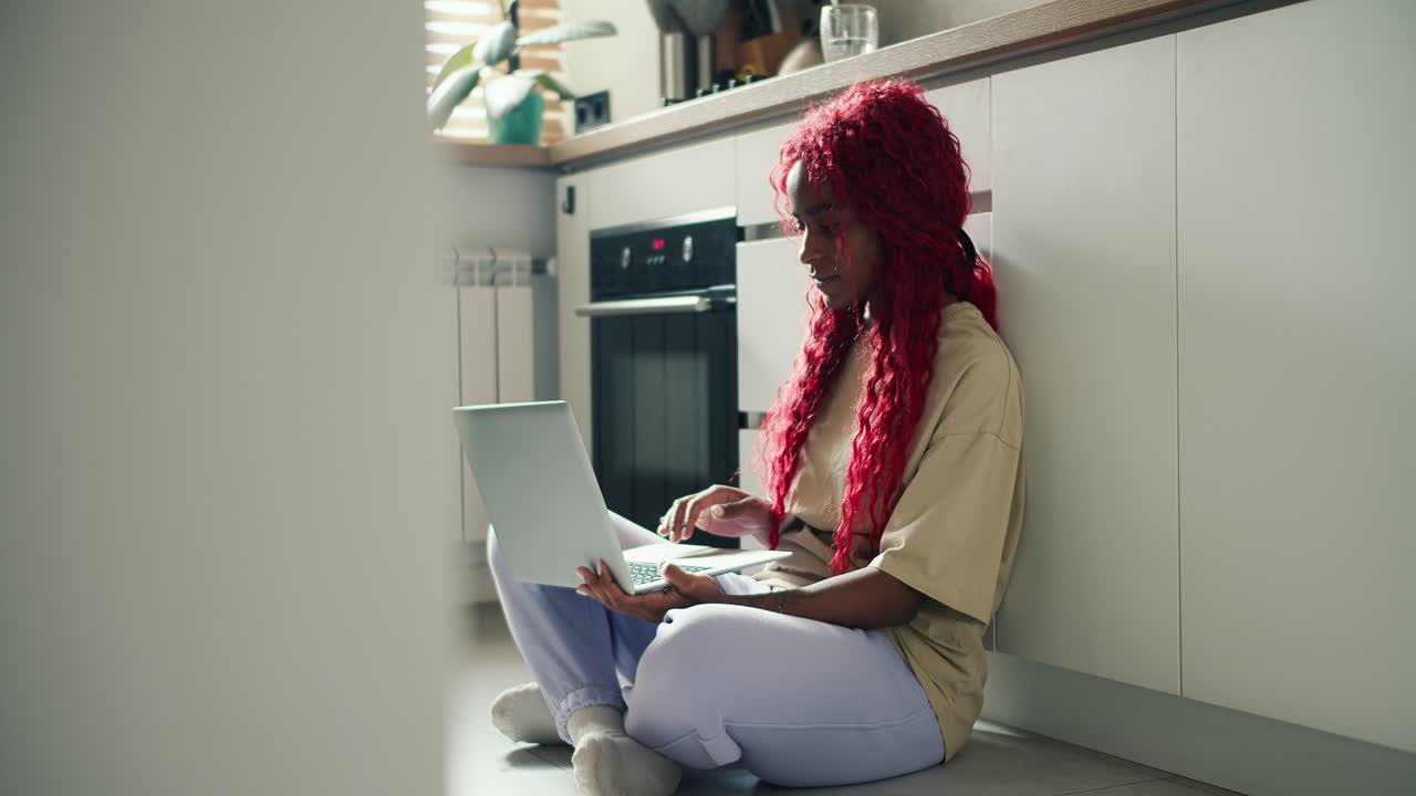 Happy astonished African American female freelancer with red curly hair sitting on kitchen floor with laptop